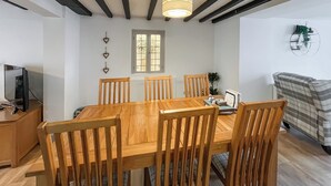 A dining area with a wooden table and chairs, a hanging light fixture, a window with blinds, and a couch in the background. - 4 bedroom accommodation in Beaumaris (Beaumaris)