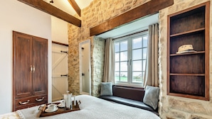Une chambre avec un mur en pierre, des poutres apparentes, une grande fenêtre avec des rideaux, une armoire en bois et une table de chevet avec un plateau.