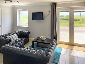 Ein Wohnzimmer mit schwarzem Ledersofa, einem wandmontierten Flachbildfernseher, einem kleinen Tisch mit einer Schüssel und Blick auf eine grüne Landschaft durch eine Schiebetür.
