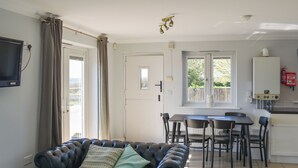 Ein Wohnzimmer mit Ledersofa, einem Esstisch mit Stühlen, einer Küchenzeile inklusive Kühlschrank und einem an der Wand befestigten Fernseher.