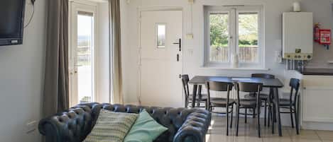 Ein Wohnzimmer mit Ledersofa, einem Esstisch mit Stühlen, einer Küchenzeile inklusive Kühlschrank und einem an der Wand befestigten Fernseher.