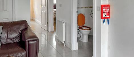 Una habitación con un sofá de cuero marrón, una mesa de centro de madera, un radiador blanco, un inodoro blanco con asiento de madera y una manta ignífuga roja en la pared.