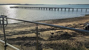 Una platja amb un moll que s'endinsa en aigües tranquil·les, una barana metàl·lica i un cel serè.