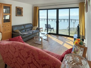 Uma sala de estar com sofá, mesa de centro de vidro e vista para o mar através de grandes janelas.