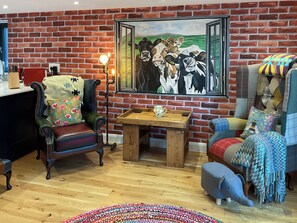A cozy room with a brick wall, a painting of cows, a wooden table, and a plush elephant chair.