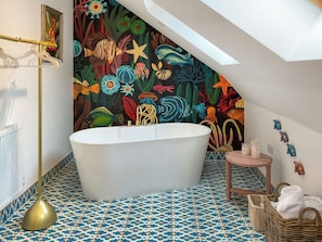 A bathroom with a freestanding bathtub, a wall mural of marine life, patterned floor tiles, and a small wicker side table with a vase. - Carreg Gwenlais Barn (Llandybie Ammanford, near Llandeilo)