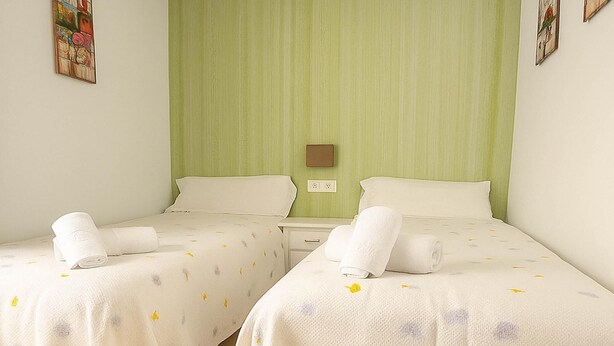 Ein Hotelzimmer mit zwei Betten, jeweils mit weißer Bettdecke und dezentem Muster. Auf jedem Bett liegen gerollte Handtücher, und die Wand des Zimmers ist grün mit einem kleinen Bilderrahmen.