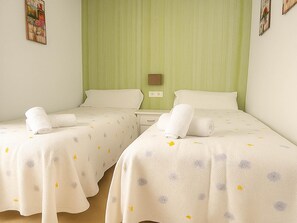 A hotel room with two beds, each with a white bedspread featuring a subtle pattern. There are two rolled towels on each bed, and the room has a green wall with a small picture frame. - Bungalow Cristina in Estepona (Estepona)