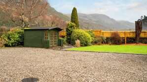A green wooden structure with a door, surrounded by gravel, a hedge, and a wooden fence.