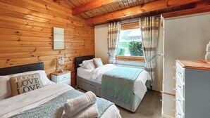 Quarto aconchegante com paredes de madeira, duas camas, uma mesa de cabeceira, cômoda e janela com cortinas.