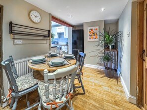 Un comedor con una mesa redonda dispuesta para cuatro, sillas con cojines estampados, un reloj de pared y una planta en maceta.