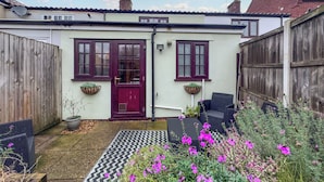 Ein Haus mit roter Tür, zwei Fenster mit Rollläden, einem Holzzaun und einem Garten mit violetten Blumen.