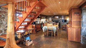 Ein rustikaler Innenraum mit einer Holztreppe, Steinwänden, einem Essbereich mit Tisch und Stühlen sowie einer Küche mit Holzausstattung.