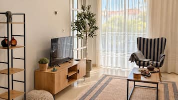 Uma sala de estar moderna com televisão, uma poltrona listrada, uma janela grande com cortinas, uma planta e um armário de madeira.