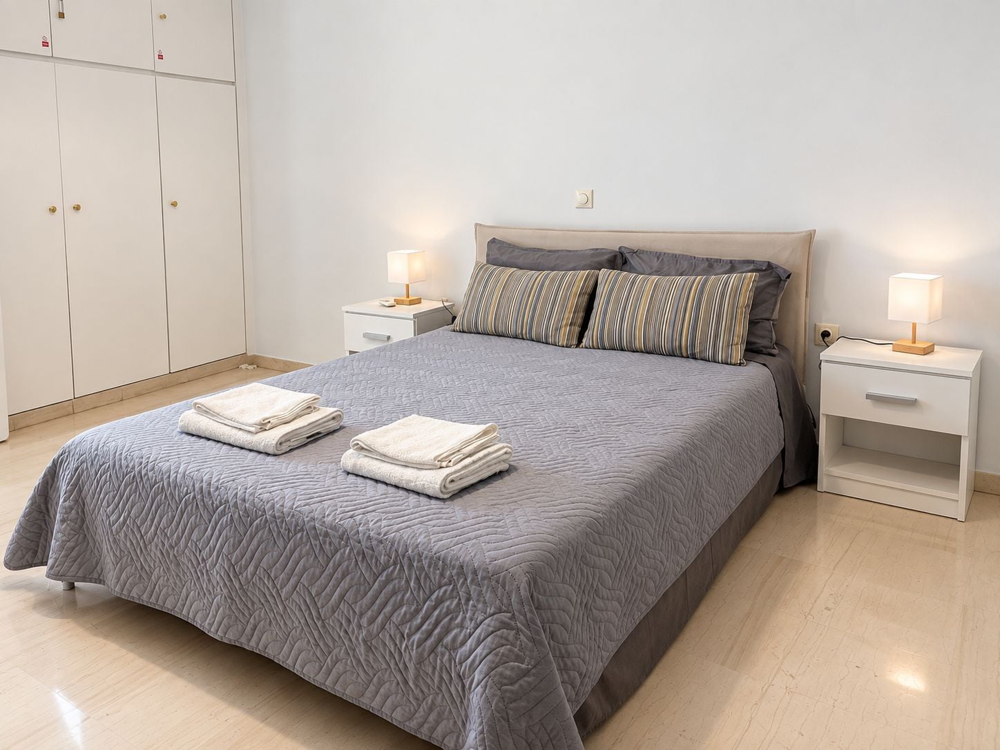 A neatly made bed with a grey quilt, two bedside tables with lamps, and a wardrobe with multiple doors.