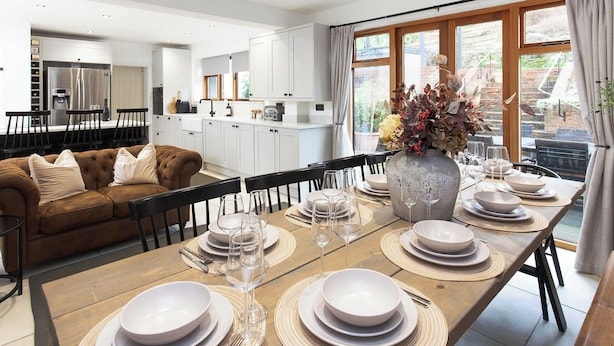 Una cocina moderna con armarios blancos, una zona de comedor con una mesa grande dispuesta para seis personas, y un sofá marrón acogedor.