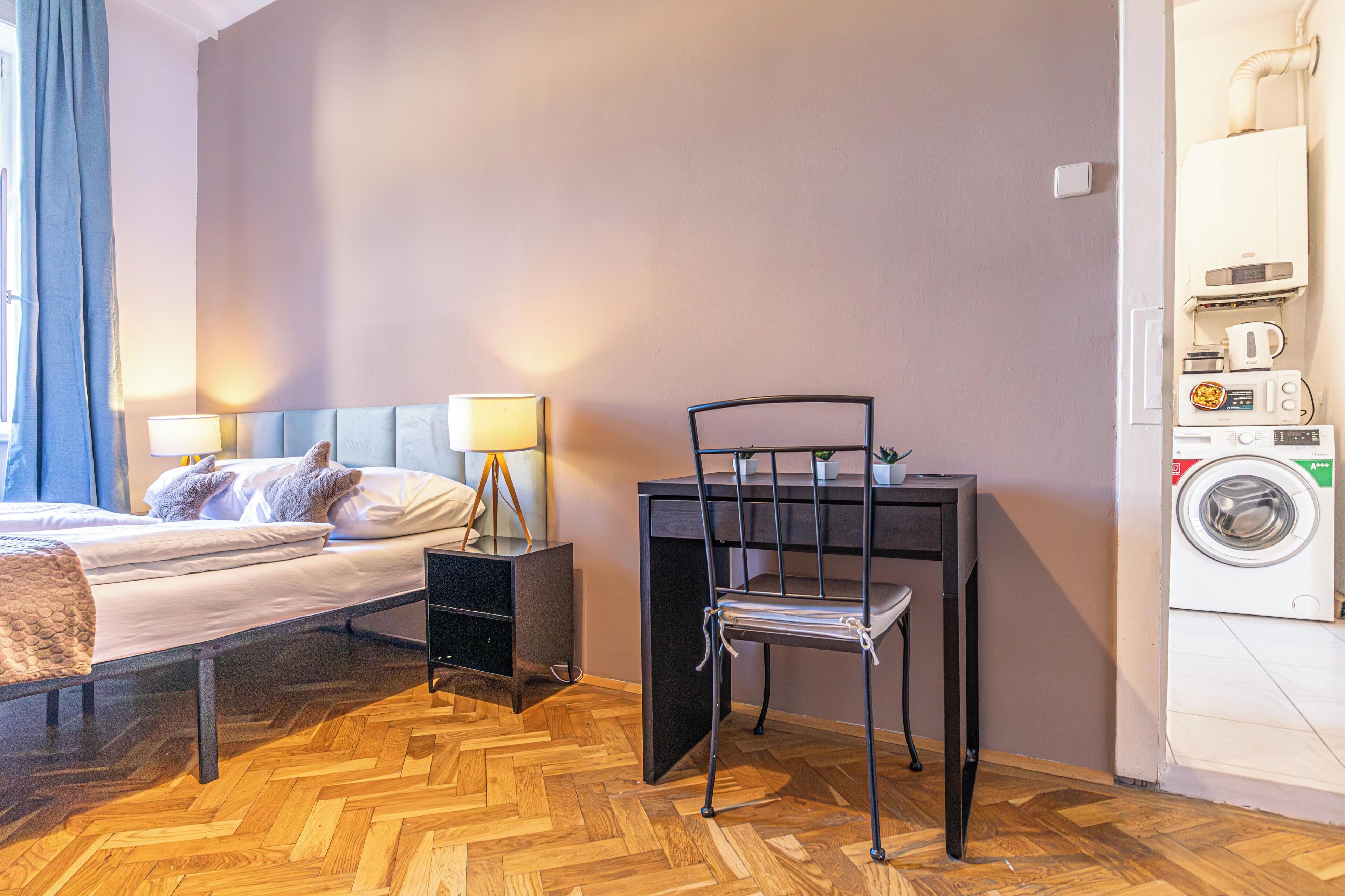 A compact hotel room with a bed, bedside lamps, a small table with a chair, a washing machine, and a herringbone wooden floor.