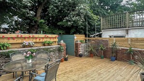 A wooden deck with a round table and chairs, surrounded by potted plants and a brick wall. - Ocean Breeze - UK47847 (Paignton)