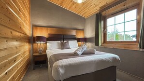 Et soveværelse med træloft, en stor seng med puder og dyne, to sengeborde med lamper, samt et vindue med gardiner.