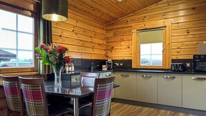 Interior de una cabaña de madera con una mesa de comedor, sillas, una zona de cocina y un ventanal grande.