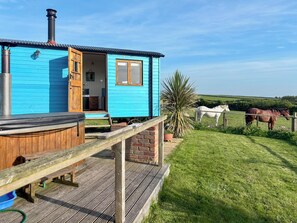 Een blauw houten huis met een houten terras, een jacuzzi en uitzicht op paarden op de achtergrond.