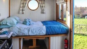 Uma cabana compacta com uma cama de solteiro, estrutura de madeira, uma pequena janela com cortinas e vista para uma área externa gramada.