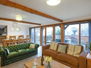 Ein geräumiges Wohnzimmer mit einem grünen Ledersofa, einem braunen Ledersofa und einem Couchtisch aus Holz. Es gibt eine Essecke mit Holztisch und -stühlen, und im Hintergrund eine Küche.