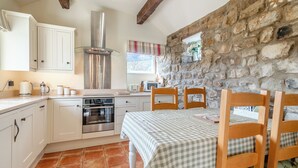 Eine Küche mit weißen Schränken, einem Herd und einer Steinwand. Ein Tisch mit karierter Tischdecke und Holzstühlen steht dort.