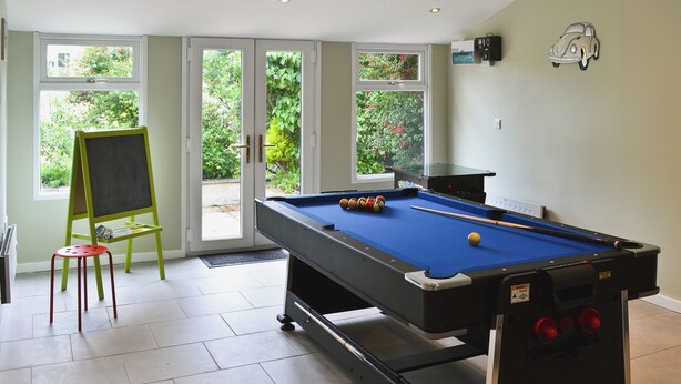 Une table de billard dans une pièce où se trouvent un fauteuil vert, un tableau noir et une vue sur l’extérieur à travers des portes vitrées.
