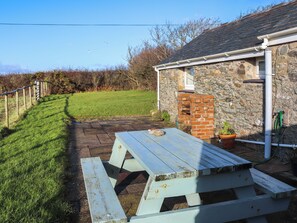 A stone building with a brick chimney, a wooden picnic table, and a grassy area with a fence. - 1 bedroom accommodation in Llanfaethlu, near Church Bay (Llanfaethlu, near Church Bay)