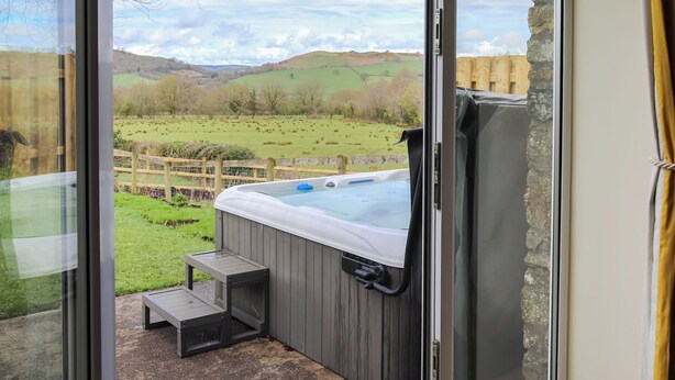 Un bain à remous avec une terrasse en bois et des marches, un mur en pierre et une vue sur un champ verdoyant et des collines.