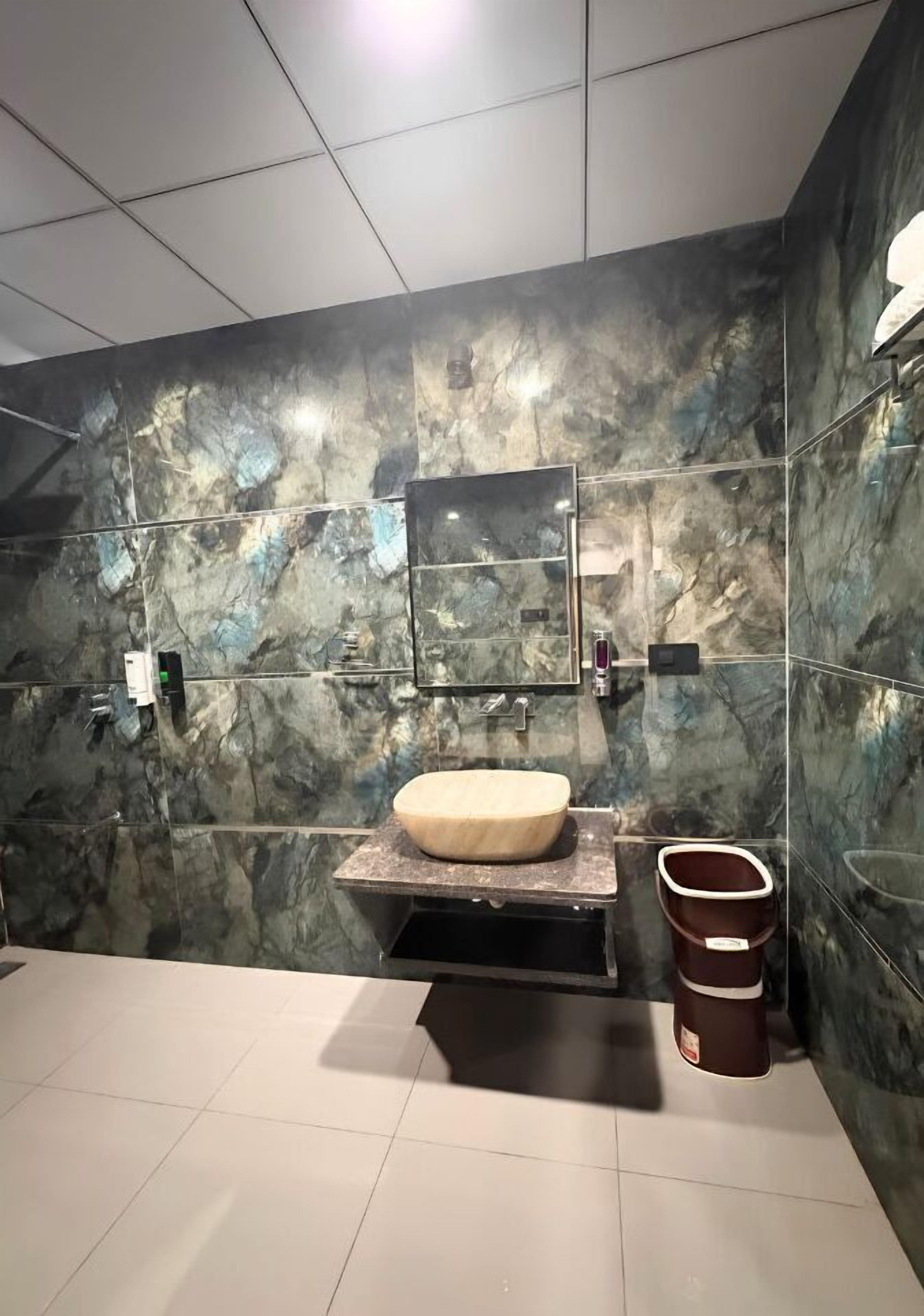 A bathroom with a marble wall, a wooden vanity, a mirror, a toilet, and a trash bin.