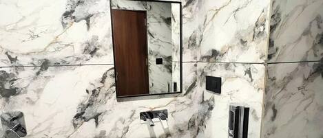 A bathroom with a marble wall, a sink, a mirror, and a shower area.