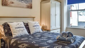 A bedroom with a bed featuring a dark blue bedspread with white scallop shell patterns, two pillows with 'Cornwall is my happy place' text, a wardrobe, and a window with a view of buildings. - 2 bedroom accommodation in Looe (Looe)