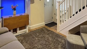 A living room with a grey sofa, a wooden console table with a vase of flowers, a flat-screen TV mounted on the wall, and a staircase leading to another room. - 2 bedroom accommodation in Looe (Looe)