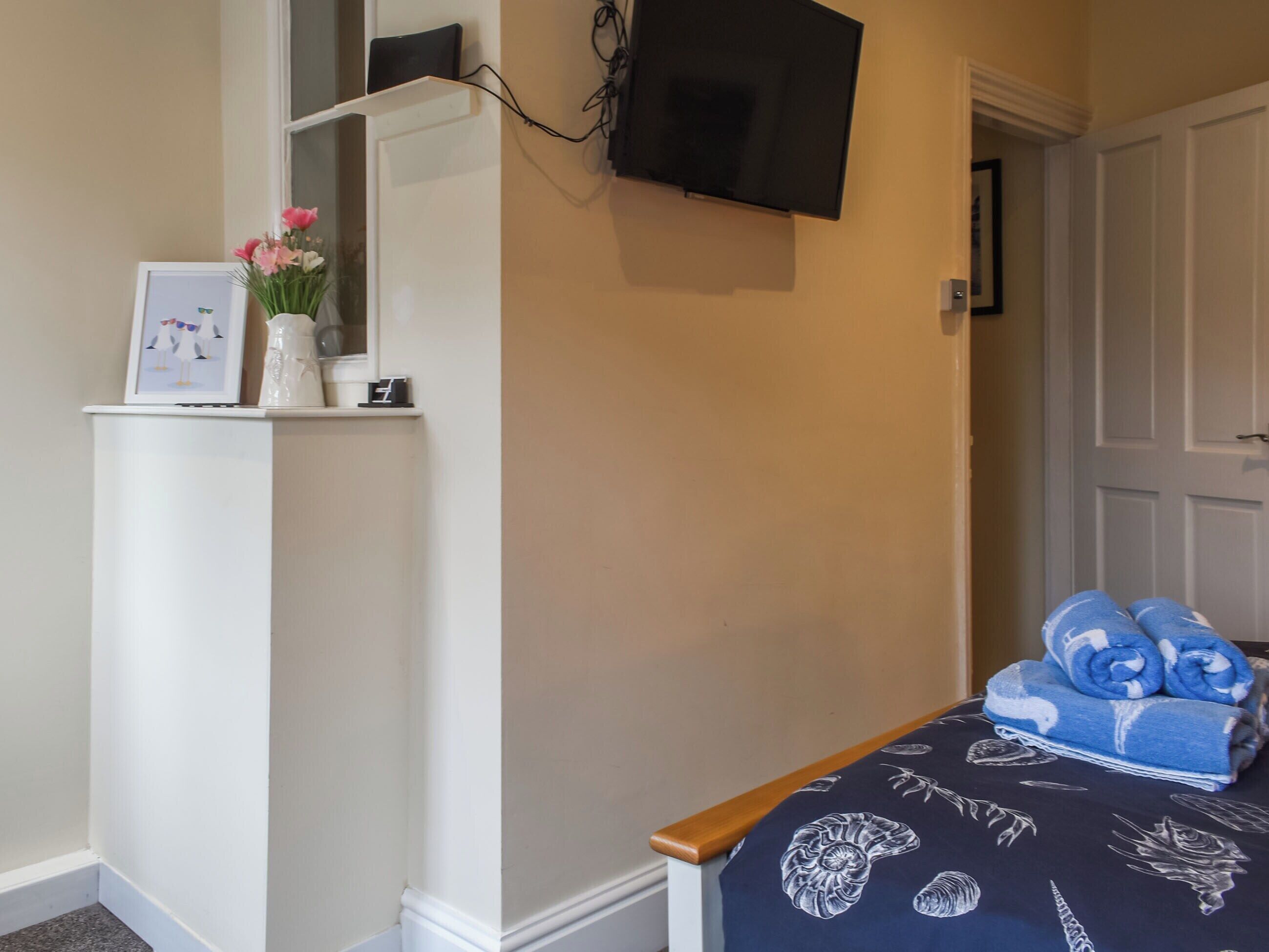 A bedroom with a bed, a television mounted on the wall, a shelf with decorative items, and a door leading to another room.