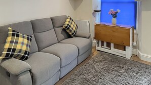 A living room with a grey sofa, plaid cushions, a small wooden side table with a vase, a blue wall, and a television. - 2 bedroom accommodation in Looe (Looe)