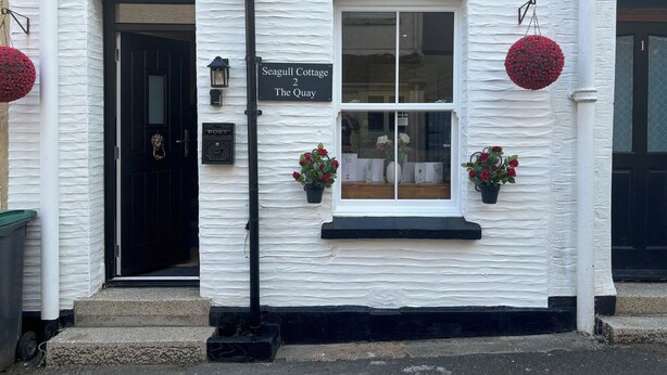 A white building with a black door, a sign reading "Seagull Cottage 2 The Quay", and two flower pots on the windowsill. - 2 bedroom accommodation in Looe (Looe)