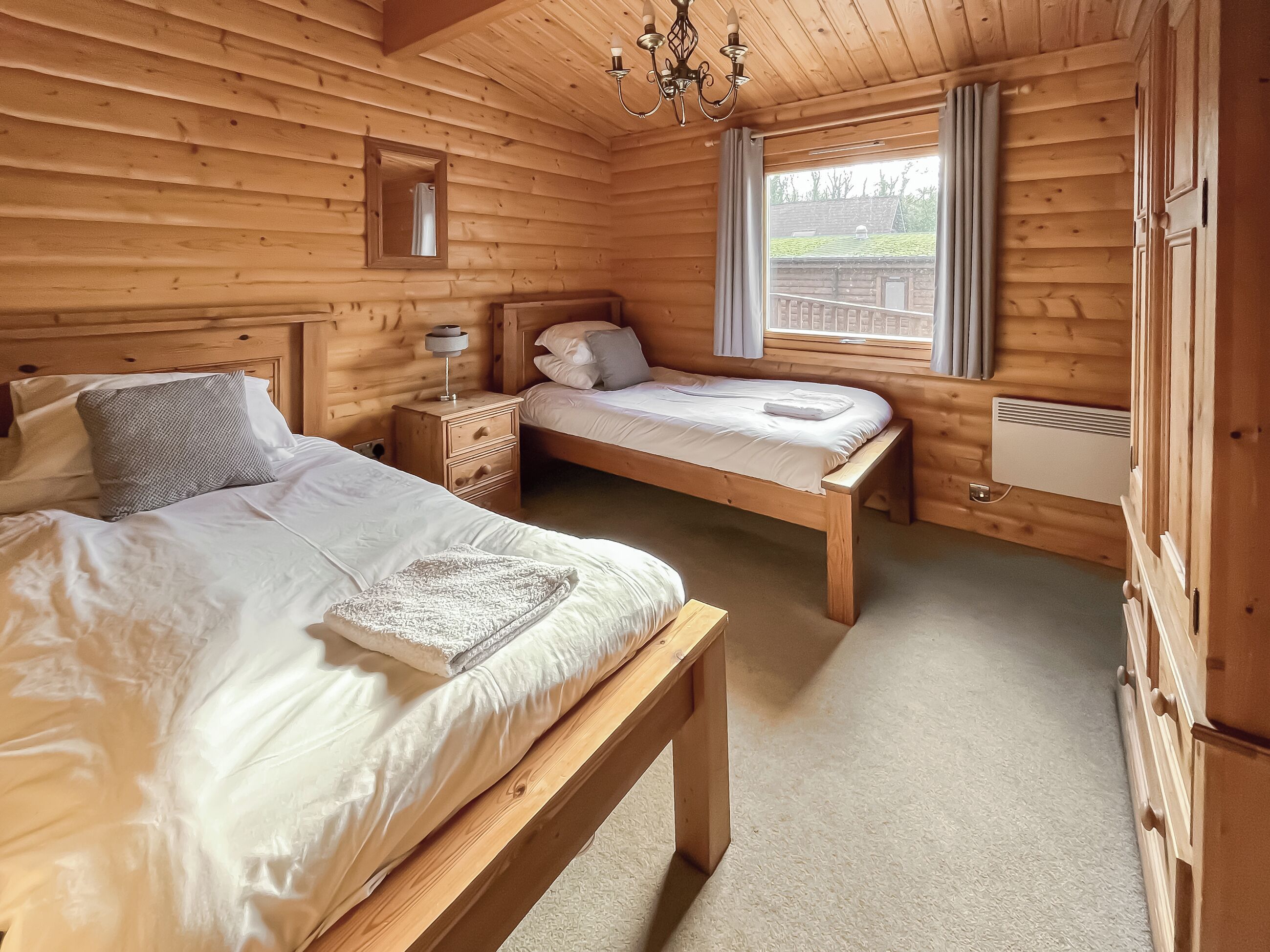A wooden cabin bedroom with two beds, a nightstand, a bench, a window with curtains, and a ceiling light.