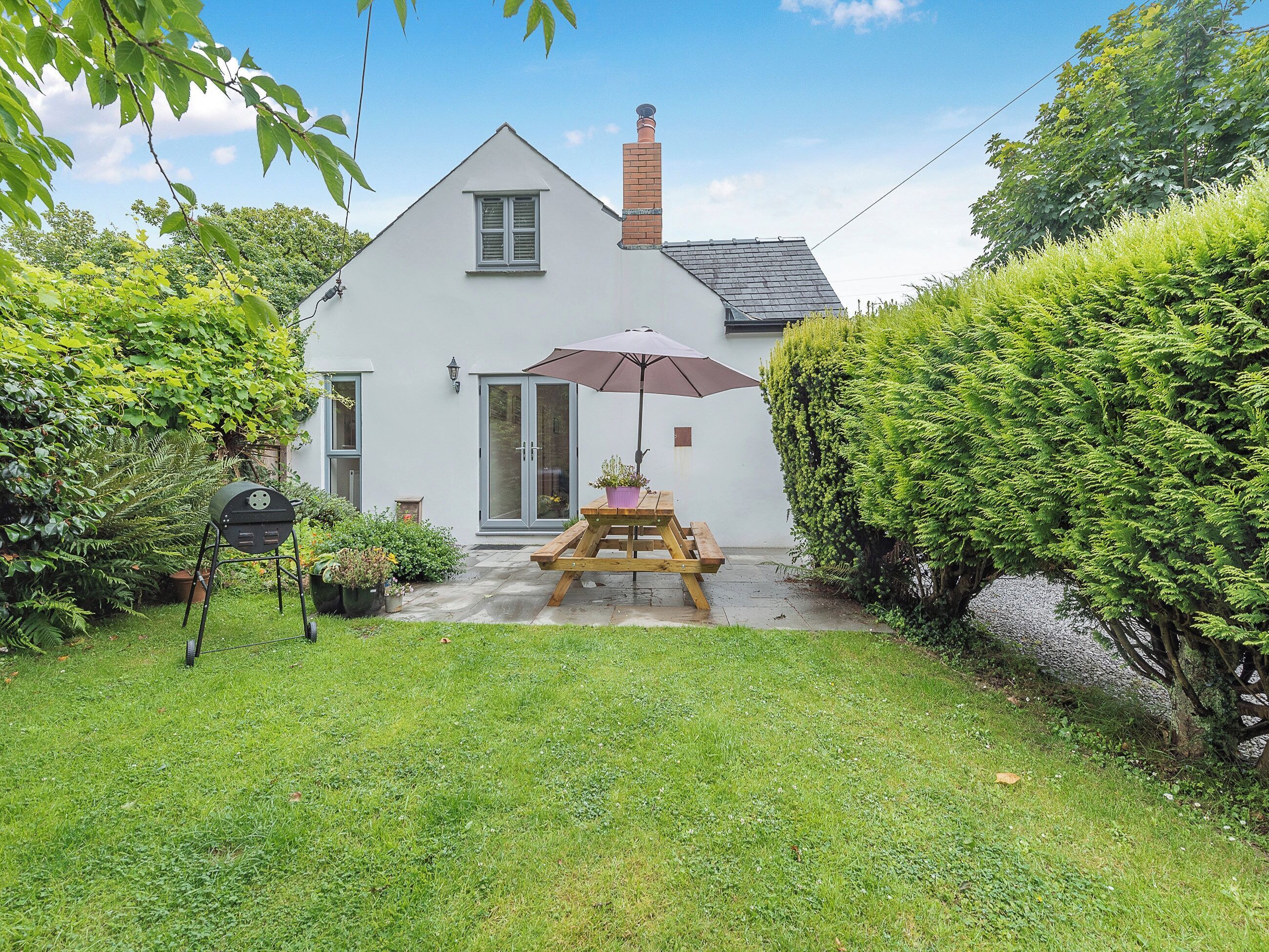 Une maison blanche avec une cheminée, un banc en bois et une table avec un parasol dans le jardin.