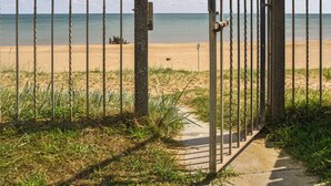 Ein Metallgitter führt zu einem Sandstrand bei blauem Himmel.