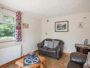 A cozy living room with a brown leather sofa, a wooden coffee table with a board game, a window with floral curtains, and a framed picture on the wall.