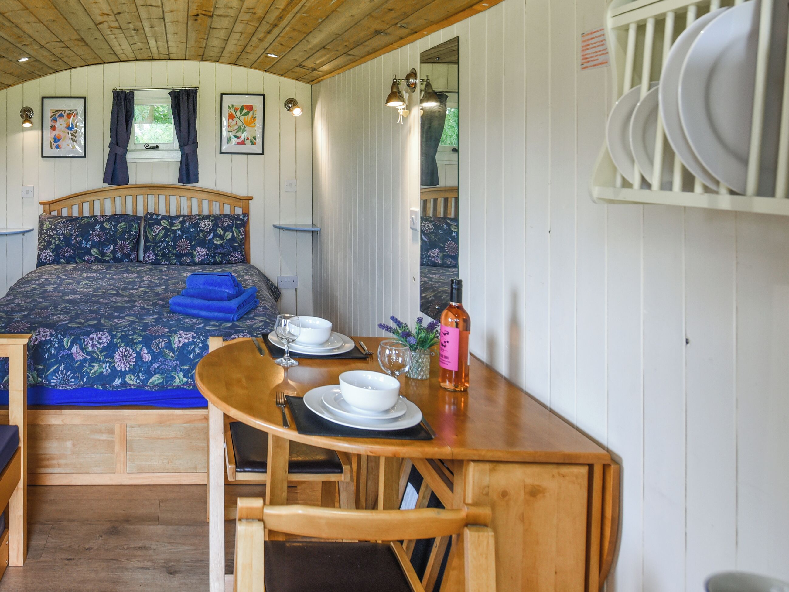 Een houten bed met bloemenbedekking, een houten tafel met een schaal, een wijnglas en een fles, en een stoel.