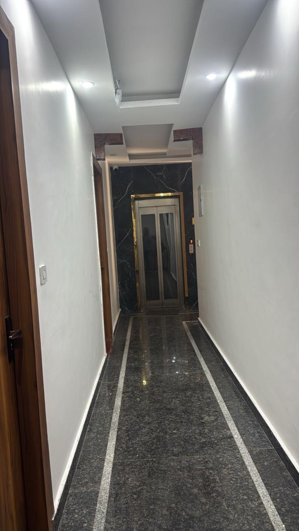 Un pasillo con piso de mármol, una puerta a la izquierda y un ascensor en el centro.