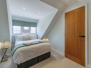 A bedroom with a bed, a window with patterned curtains, a wooden door, and a small bedside table with a lamp.