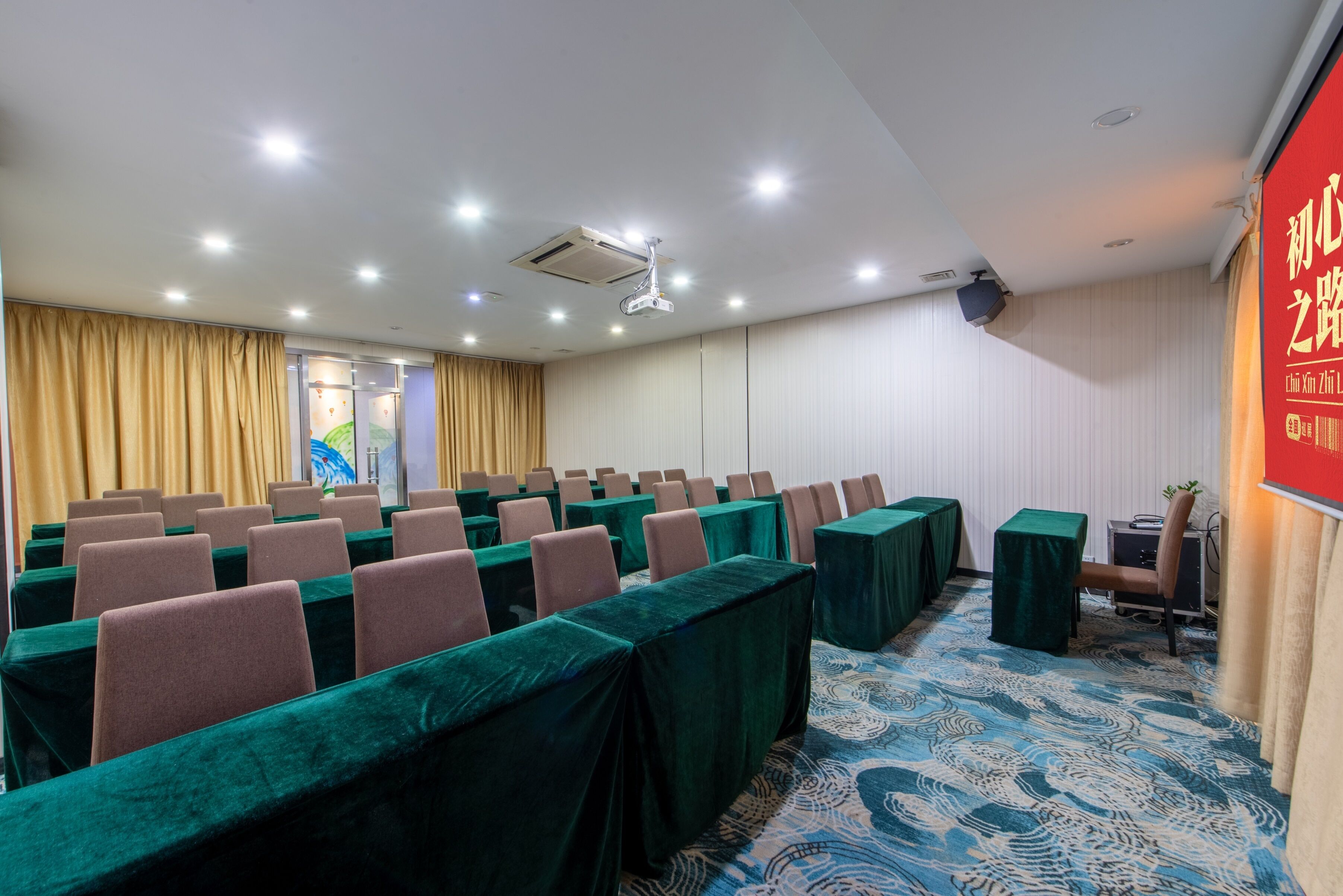 Un salón de conferencias con filas de mesas y sillas, un proyector y un cartel rojo en la pared.