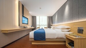 Een moderne hotelkamer met een groot bed, een aan de muur bevestigde televisie, een houten nachtkastje met een telefoon en een tijdschrift, en een houten vloer.