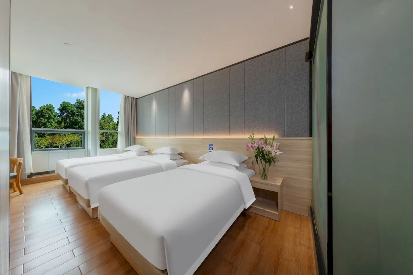 Habitación de hotel moderna con dos camas, piso de madera, un ventanal con vistas a árboles y una mesita de noche con un jarrón de flores.