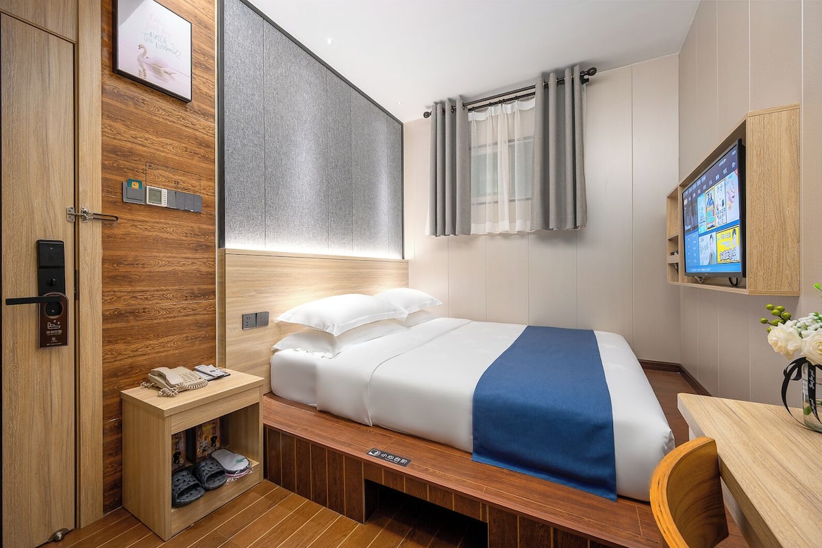Une chambre d’hôtel moderne avec une armoire en bois, un lit avec une literie blanche et bleue, une table de chevet avec un téléphone et un téléviseur fixé au mur.