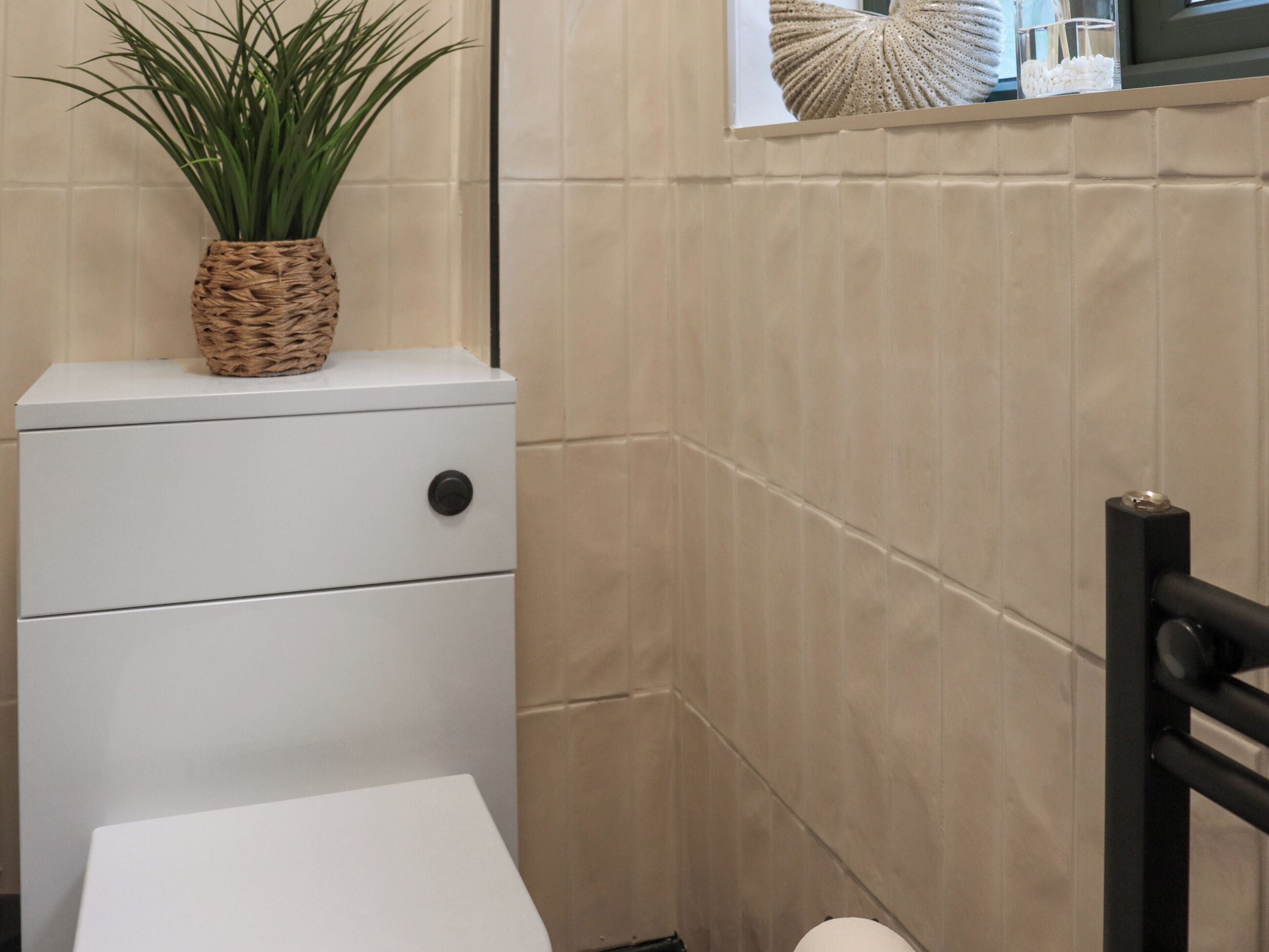 A bathroom with a white toilet, a wicker plant on top, and a tiled wall.